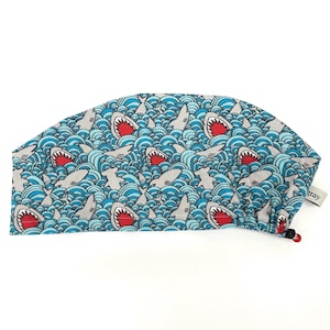 May include: A blue surgical cap with a repeating pattern of gray sharks with red mouths swimming in blue and white waves. The cap has a tag that says "gray".