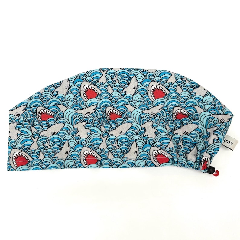 May include: A blue surgical cap with a repeating pattern of gray sharks with red mouths swimming in blue and white waves. The cap has a tag that says "gray".