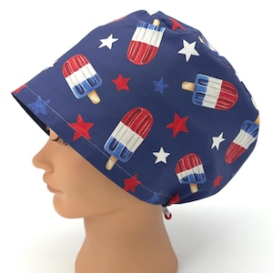 May include: A navy blue scrub cap featuring a repeating pattern of red, white, and blue popsicle illustrations interspersed with solid red and white star shapes. The cap has a rounded crown and a tie-back closure with a small red toggle at the base.