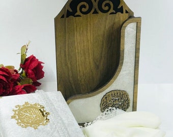 May include: A white book with gold accents and a decorative gold emblem, a small digital counter, and a gold tassel with white flowers. A wooden holder with a decorative cutout design and a white fabric with lace trim are also present.