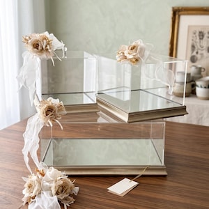 May include: Three clear acrylic display boxes with mirrored bases and metallic gold-toned trim. Each box is adorned with decorative fabric flowers in shades of cream and beige, featuring flowing white ribbons and a small blank rectangular tag.