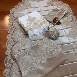 May include: An elegant prayer mat set in cream and beige, featuring intricate gold lace detailing. The set includes a prayer mat, a towel with fringe, a small bag, and a round pouch, all with matching ornate designs. The items are arranged on a wooden surface.
