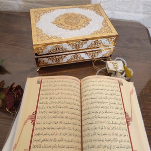Może przedstawiać: Dekoracyjne pudełko w kolorze białym i złotym z misternymi wzorami i kaligrafią arabską. Pudełko jest otwarte, a w nim widoczna jest książka z tekstem arabskim. Książka jest otwarta na stronie z czerwoną zakładką.