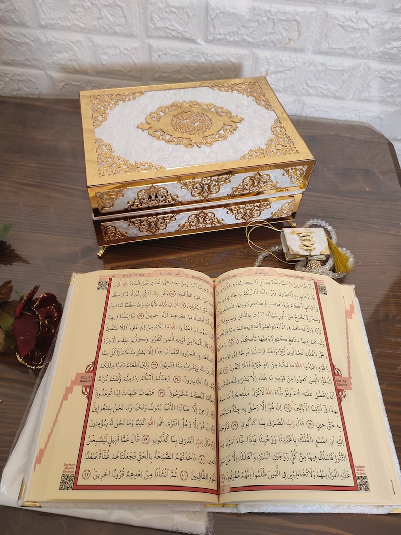 Może przedstawiać: Dekoracyjne pudełko w kolorze białym i złotym z misternymi wzorami i kaligrafią arabską. Pudełko jest otwarte, a w nim widoczna jest książka z tekstem arabskim. Książka jest otwarta na stronie z czerwoną zakładką.