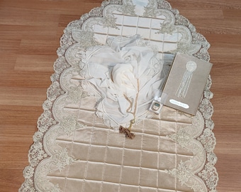 May include: A beige prayer rug with a lace border and a quilted design. The rug is decorated with a white sheer scarf and a small prayer counter. A small book with the text "Dua'a Book" is also included.