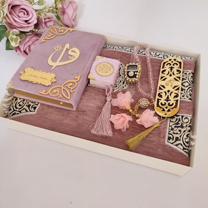 Può includere: Un set regalo malva con un tappeto da preghiera, un piccolo Corano e un libro più grande con dettagli dorati. Il set include una collana di perline, un segnalibro decorativo e decorazioni floreali rosa, il tutto in una scatola bianca.