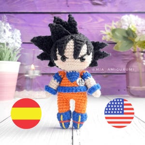 Puede incluir: Figura amigurumi de crochet de un personaje de dibujos animados con cabello negro, un atuendo azul y naranja, y el carácter japonés para "fuerza" en el pecho. La figura está de pie sobre una superficie blanca con una bandera española y una bandera estadounidense a cada lado.