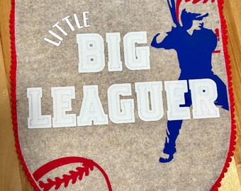 May include: Gray felt pennant with red trim and a baseball theme. The pennant features the words "LITTLE BIG LEAGUER" in white letters, a baseball illustration, and a blue baseball player silhouette.