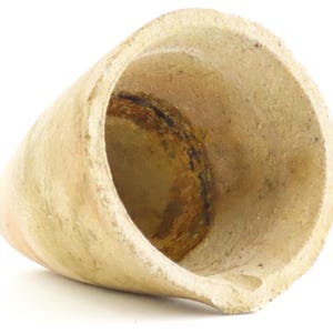 May include: A weathered, cone-shaped ceramic vessel with a wide opening and a rounded base. The interior shows dark staining, and the exterior has a light beige color. The vessel's shape suggests it may have been used for holding liquids or solids.