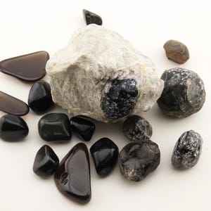May include: A collection of various stones including a large, rough, light gray rock with a dark embedded crystal, several smooth, polished black stones, translucent dark brown pieces, and textured, dark gray mineral specimens.