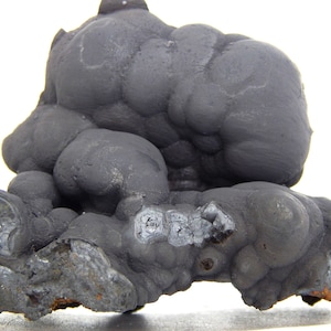 May include: A large, dark gray, botryoidal mineral specimen with a rough, textured surface. The mineral has a rounded, bubbly appearance and is set against a white background.