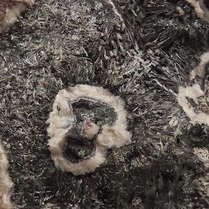 May include: A mineral specimen featuring a dense cluster of metallic, dark gray, needle-like crystals interspersed with patches of opaque, off-white, and light brown mineral deposits, creating a textured, rugged surface with distinct circular patterns.