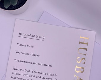 Puede incluir: Una tarjeta blanca con letras doradas que dicen "Husband". La tarjeta tiene una definición de la palabra "husband" con una cita sobre el fruto de la boca de un hombre. La tarjeta está sobre una superficie blanca con una pequeña planta suculenta verde en una maceta blanca.