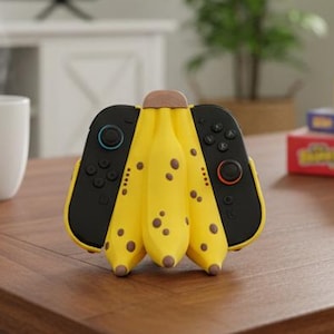 May include: A unique yellow banana-shaped video game controller with black buttons and a brown tip sits on a wooden coffee table. The controller is next to a white mug and a television playing a racing game.