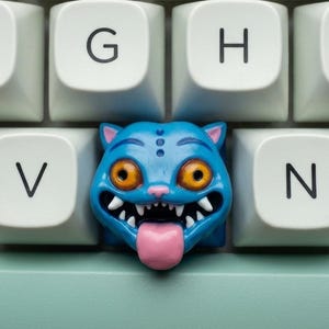 May include: A close-up of a keyboard with white keys and a custom blue cat-shaped keycap. The cat keycap has orange eyes, a pink tongue, and sharp white teeth. The keyboard has a light green base.