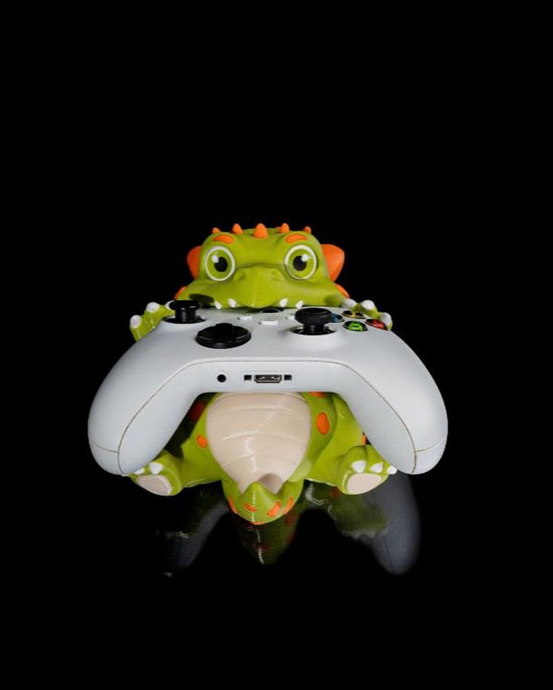 Puede incluir: Un soporte para mando con forma de dinosaurio verde y naranja que sostiene un mando Xbox blanco. El soporte tiene p&uacute;as y manchas naranjas. El mando est&aacute; centrado, mostrando sus botones y puertos.