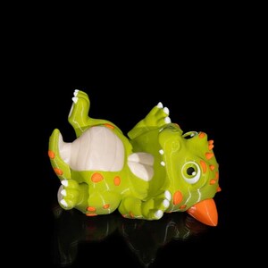 Puede incluir: Una figura de dinosaurio verde y naranja tumbada boca arriba. La figura tiene manchas naranjas, una barriga color crema y un pico naranja. Los ojos del dinosaurio son grandes y redondos, con pupilas negras.
