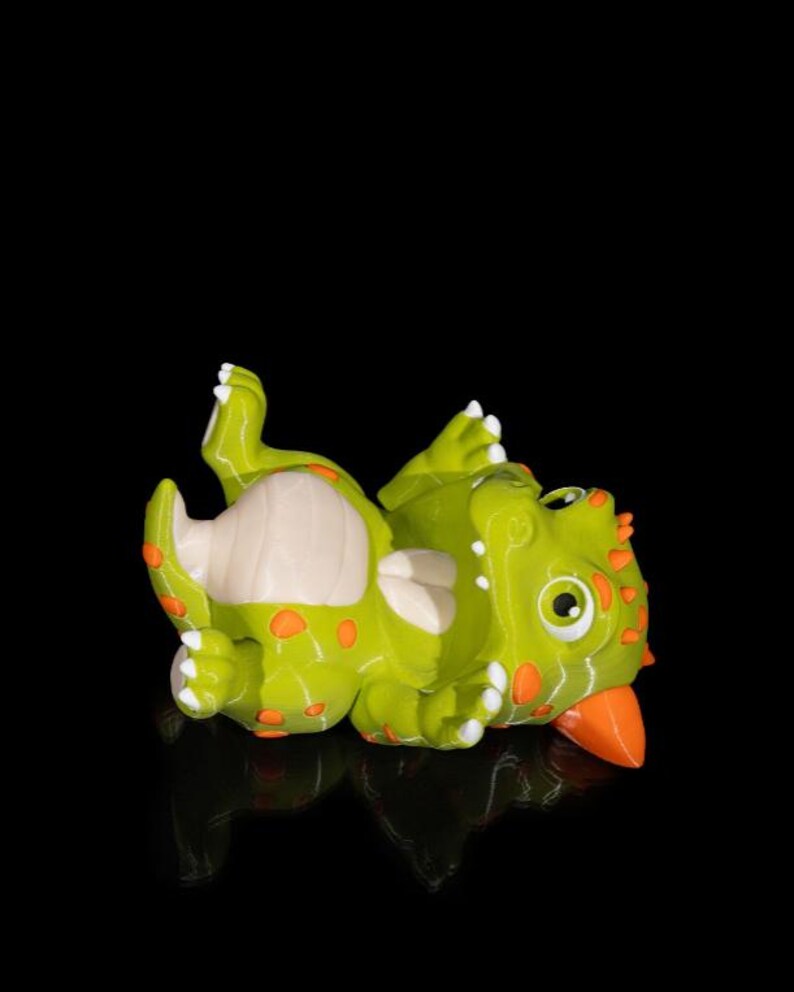 Puede incluir: Una figura de dinosaurio verde y naranja tumbada boca arriba. La figura tiene manchas naranjas, una barriga color crema y un pico naranja. Los ojos del dinosaurio son grandes y redondos, con pupilas negras.