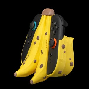 K&ouml;nnte beinhalten: Ein neuartiges, bananenf&ouml;rmiges Zubeh&ouml;r f&uuml;r eine Spielkonsole. Das gelbe Zubeh&ouml;r hat braune Flecken und einen braunen Stiel, mit schwarzen Gamecontrollern auf beiden Seiten. Der Hintergrund ist einfarbig schwarz.