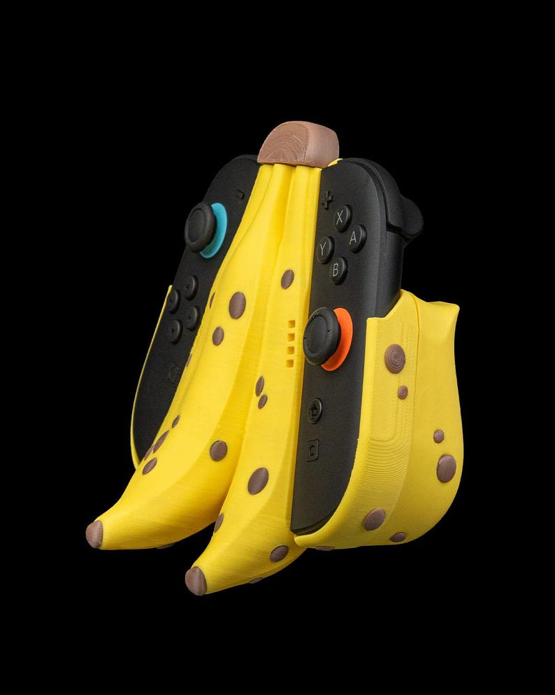 K&ouml;nnte beinhalten: Ein neuartiges, bananenf&ouml;rmiges Zubeh&ouml;r f&uuml;r eine Spielkonsole. Das gelbe Zubeh&ouml;r hat braune Flecken und einen braunen Stiel, mit schwarzen Gamecontrollern auf beiden Seiten. Der Hintergrund ist einfarbig schwarz.
