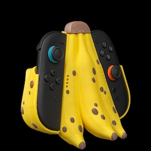 K&ouml;nnte beinhalten: Ein skurriler, bananenf&ouml;rmiger Halter f&uuml;r Nintendo Switch Joy-Con-Controller. Der gelbe Halter hat braune Flecken und eine Holzoberseite, mit zwei schwarzen Controllern. Der Hintergrund ist schwarz.