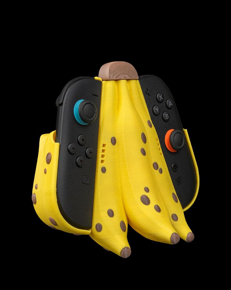 K&ouml;nnte beinhalten: Ein skurriler, bananenf&ouml;rmiger Halter f&uuml;r Nintendo Switch Joy-Con-Controller. Der gelbe Halter hat braune Flecken und eine Holzoberseite, mit zwei schwarzen Controllern. Der Hintergrund ist schwarz.