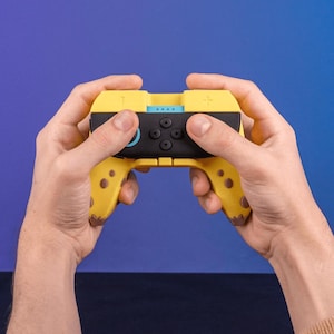 May include: A yellow and black video game controller with a blue accent. The controller has a black centre with four black buttons and a directional pad. The handles have a dotted pattern. The controller is held by two hands against a blue background.