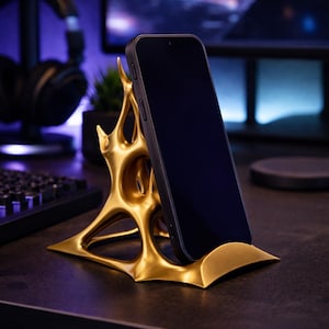 Könnte beinhalten: Ein goldfarbener Telefonständer mit einem abstrakten, organischen Design. Ein schwarzes Smartphone befindet sich in dem Ständer. Der Ständer hat eine breite Basis und eine komplexe, durchbrochene Struktur, die das Telefon hält. Der Hintergrund ist unscharf.