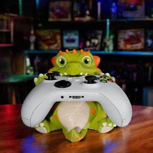 Puede incluir: Un soporte para mando con forma de dinosaurio verde y naranja que sostiene un mando de juego blanco. El soporte tiene ojos grandes y detalles en naranja. El mando es blanco con botones negros. El soporte est&aacute; sobre una superficie de madera.