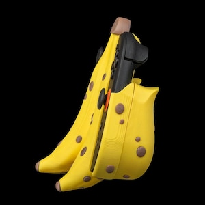 K&ouml;nnte beinhalten: Eine gelbe Bananen-f&ouml;rmige H&uuml;lle mit braunen Flecken, die ein schwarzes elektronisches Ger&auml;t enth&auml;lt. Die H&uuml;lle hat eine braune Spitze und ist vor einem schwarzen Hintergrund platziert. Das Ger&auml;t hat schwarze Kn&ouml;pfe und einen orangefarbenen Akzent.