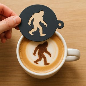 May include: A black coffee stencil with a Bigfoot silhouette is held above a white coffee mug with a Bigfoot design in the foam. The stencil is round with a small handle. The coffee has a creamy, light brown color.