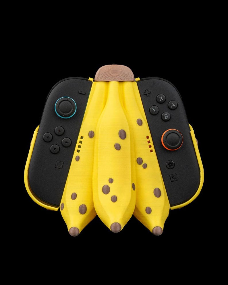 K&ouml;nnte beinhalten: Ein einzigartiges, gelbes, bananenf&ouml;rmiges Zubeh&ouml;r f&uuml;r eine Spielkonsole mit braunen Flecken und einer Holzspitze. Schwarze Controller mit farbigen Kn&ouml;pfen befinden sich auf beiden Seiten. Der Hintergrund ist schwarz.