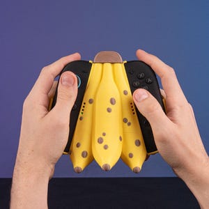 K&ouml;nnte beinhalten: Ein gelber, bananenf&ouml;rmiger Videospiel-Controller mit braunen Flecken und einem Holzaufsatz. Der Controller hat schwarze Seiten mit Kn&ouml;pfen und Joysticks. Er wird von zwei H&auml;nden vor blauem Hintergrund gehalten.