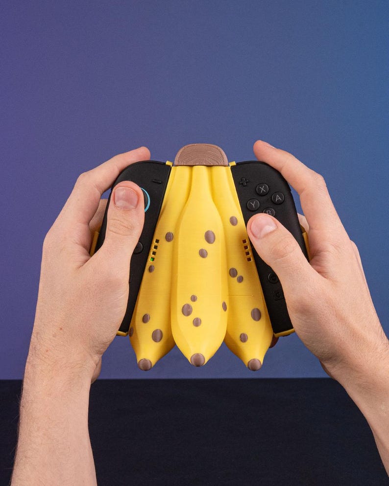 K&ouml;nnte beinhalten: Ein gelber, bananenf&ouml;rmiger Videospiel-Controller mit braunen Flecken und einem Holzaufsatz. Der Controller hat schwarze Seiten mit Kn&ouml;pfen und Joysticks. Er wird von zwei H&auml;nden vor blauem Hintergrund gehalten.