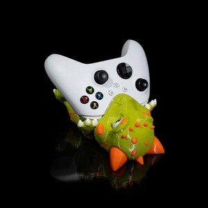 Puede incluir: Un mando Xbox blanco descansa sobre un soporte con forma de dinosaurio verde y naranja. El mando tiene botones negros y botones de acci&oacute;n de colores. El soporte tiene p&uacute;as naranjas y una textura detallada.