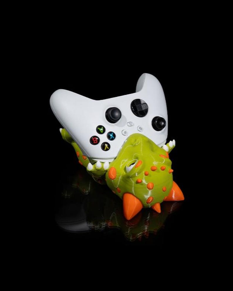 Puede incluir: Un mando Xbox blanco descansa sobre un soporte con forma de dinosaurio verde y naranja. El mando tiene botones negros y botones de acci&oacute;n de colores. El soporte tiene p&uacute;as naranjas y una textura detallada.