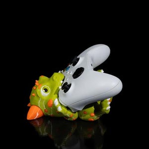 Puede incluir: Un soporte para mando con forma de dinosaurio verde y naranja que sostiene un mando de videojuegos blanco. El soporte tiene detalles naranjas y est&aacute; sobre una superficie negra reflectante.