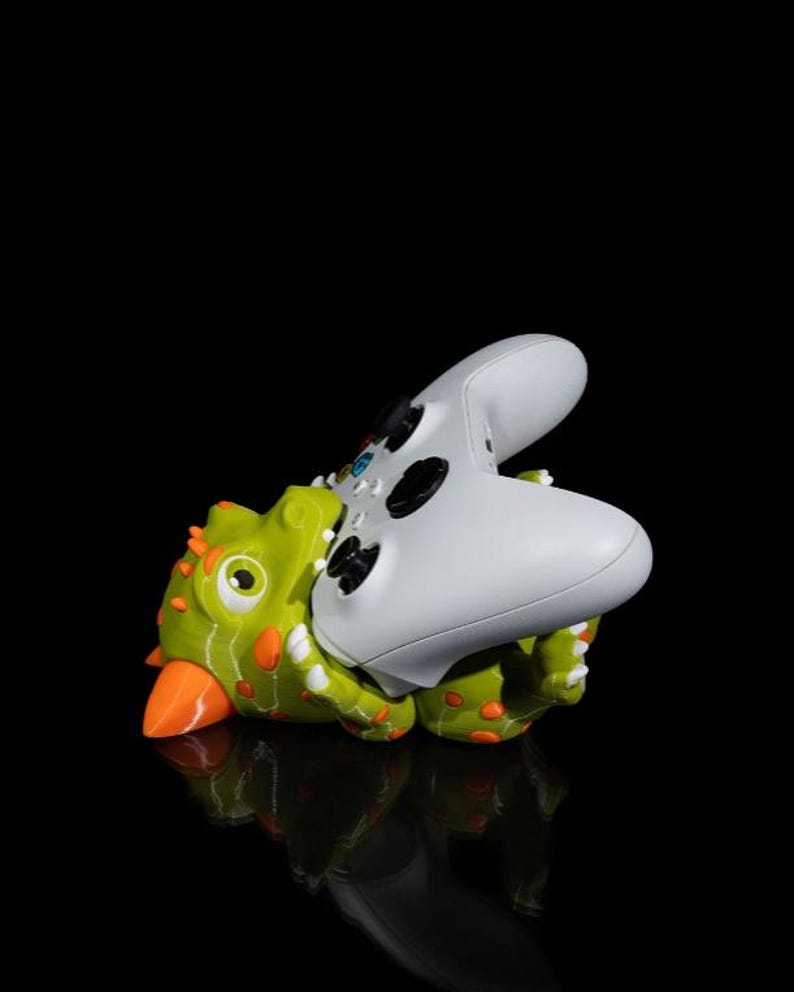 Puede incluir: Un soporte para mando con forma de dinosaurio verde y naranja que sostiene un mando de videojuegos blanco. El soporte tiene detalles naranjas y est&aacute; sobre una superficie negra reflectante.