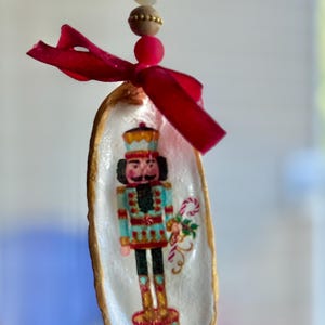 May include: A Christmas ornament featuring a painted nutcracker soldier inside a white shell with a gold rim. The ornament is adorned with a red ribbon and decorative beads, perfect for holiday decor.