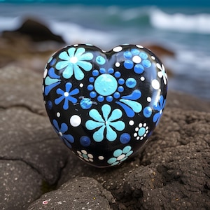 May include: A heart-shaped rock painted with a black background and blue and white floral designs. The rock is sitting on a dark gray, textured surface.