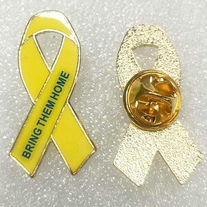 Puede incluir: Pin de cinta amarilla con las palabras "Bring Them Home" impresas en &eacute;l. El pin tiene un respaldo dorado con un pasador de seguridad.