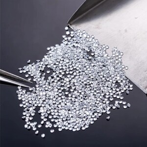 May include: A pile of small, clear, round diamonds on a black surface. The diamonds are loose and scattered, and some are spilling from a metal scoop.