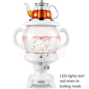 May include: White electric tea kettle with a glass teapot on top. The kettle has a spherical glass chamber with a spout and two handles. The teapot is filled with tea. The text on the image says "LED lights turn red when in boiling mode."