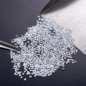 Może przedstawiać: Stosik małych, przezroczystych, okrągłych diamentów na czarnym tle. Diamenty są luźne i rozproszone, a niektóre wysypują się z metalowej łopatki.