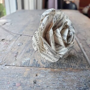 以下が含まれることがあります： 本のページで作られた紙の花。花は白く、繊細で波打った質感があります。本のページは見え、テキストは黒いインクで印刷されています。