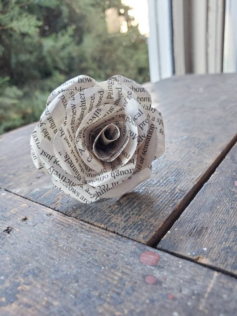 以下が含まれることがあります： 本のページから作られた紙のバラ。バラは薄い茶色で、繊細で複雑なデザインが施されています。バラは木製の表面の上に置かれています。