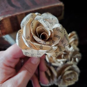 以下が含まれることがあります： 本のページで作られた、クリーム色の紙のバラが1輪。バラはカールしていて、繊細でヴィンテージな雰囲気です。バラは暗い背景の上に置かれています。