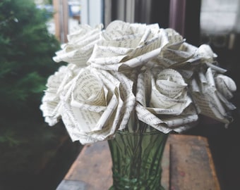以下が含まれることがあります： 緑色のガラスの花瓶に、本のページで作られた白い紙のバラのブーケ。バラは円形に配置され、花瓶は木製の表面の上に置かれている。