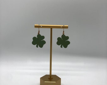May include: Olive green shamrock earrings with gold-coloured ear wires and hooks. The earrings are displayed on a gold-coloured jewellery stand with a hexagonal base. The background is plain white.