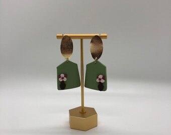 May include: A pair of green statement earrings with a gold oval top and a pink flower accent. The earrings are displayed on a gold stand.